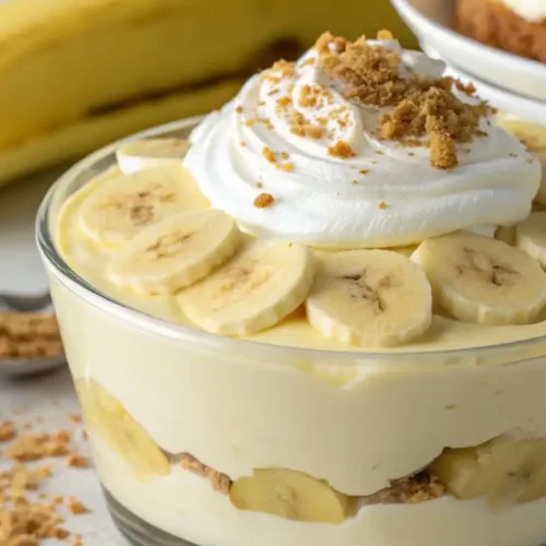 Close-up of creamy dairy-free banana pudding in a glass dish, layered with banana slices, topped with dairy-free whipped cream and crumbled graham crackers.