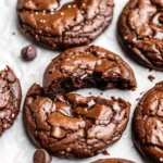 Close-up of chewy fudgy brookies — rich chocolate cookies with a glossy, crackled top, gooey chocolate chunks, and a sprinkle of sea salt on parchment paper.