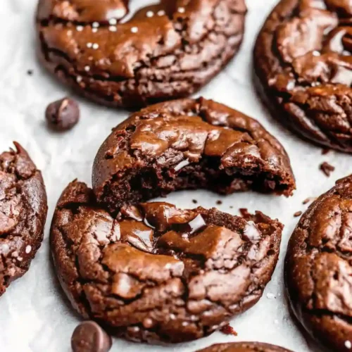Close-up of chewy fudgy brookies — rich chocolate cookies with a glossy, crackled top, gooey chocolate chunks, and a sprinkle of sea salt on parchment paper.